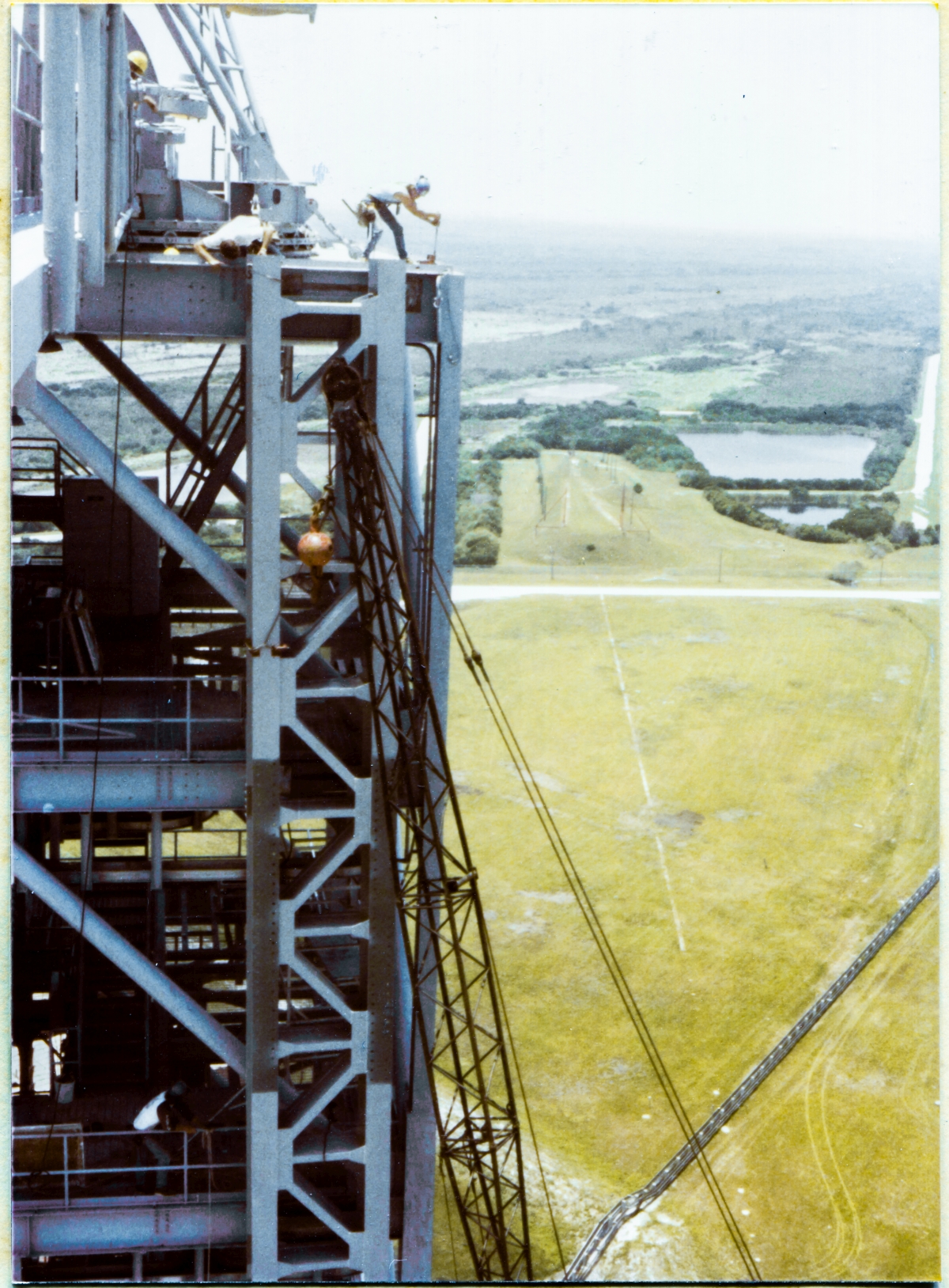 Image 079. Union Ironworker James Dixon has placed a loop of wire rope around the top of the GOX Arm Hinges Support Strongback, and is standing on top of the Fixed Service Structure at Space Shuttle Launch Complex 39-B, Kennedy Space Center, Florida, at Elevation 300'-0”, no handrails, wearing no safety harness or other gear, as was common practice in the early 1980's, working the come-along that the wire rope has been threaded to, via an unseen snatch block which is hidden behind the Strongback. Main force is now being applied, and the top of the Strongback is in hard contact with the web of the Perimeter Framing Beam of the FSS, scraping along against it as it is urged ever northward by the action of the come-along, having been shoved into that hard contact by the further action of other Union Ironworkers two levels down, at Elevation 260'-0”, who are slowly tensioning the wire rope around the second-to-bottom horizontal bracing member at the lower end of the Strongback, forcing it into a more-vertical orientation, going against its natural tendency to hang at an angle because of where its lifting sling remains attached, roughly one-third down from its top. The Strongback is not yet fully vertical and is only contacting the FSS at its top connection point. Below that, the other two connection points remain out of contact, and the Strongback is still reasonably free to be pushed side-to-side, along the face of the FSS. At top and bottom, other Union Ironworkers are in constant communication with the rest of the gang, closely-monitoring the position of the Strongback and its connection points. Photo by James MacLaren.